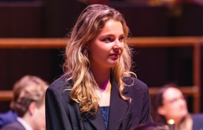 A blonde-haired woman looks to her right. She is presenting at an event, surrounded by seated attendees.