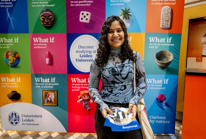 A girl with black hair wearing a black and blue top stands in front of university campaign posters saying 'What if?'