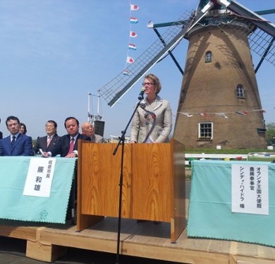 A woman with medium-length hair stands at a microphone. Behind her is a windmill.