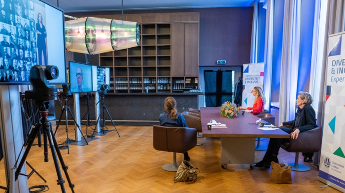 Three people sit at a table in front of a large screen. On the screen are symposium participants.