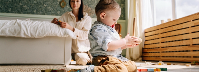 Baby with toy on the foreground, woman next to a bed in the background.