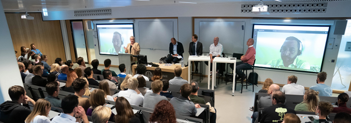 Photo of the hall where the debate took place. In the back, people are sitting in rows. In the front, four presenters and on panelist on a projector through a video call.